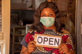 A woman wearing a mask stands in a doorway holding an "OPEN" sign.