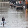 A person walking through a flooded street while carrying bags amidst high water levels, with parked vehicles partially submerged in the background. A person walking through a flooded street while carrying bags amidst high water levels, with parked vehicles partially submerged in the background.