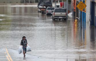 A person walking through a flooded street while carrying bags amidst high water levels, with parked vehicles partially submerged in the background.