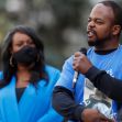 A man speaks into a microphone at a public event, while a woman in a blue outfit listens attentively in the background. A man speaks into a microphone at a public event, while a woman in a blue outfit listens attentively in the background.