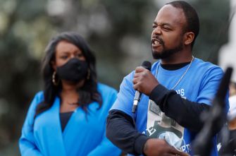 A man speaks into a microphone at a public event, while a woman in a blue outfit listens attentively in the background.