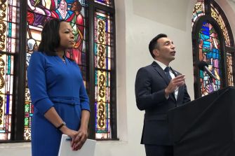 A press conference featuring two speakers, one in a blue dress and the other in a suit, standing at a podium with stained glass windows in the background.