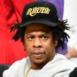 Jay-Z seated, looking contemplative, wearing a black cap with the word "RHUDE" on it. Jay-Z seated, looking contemplative, wearing a black cap with the word "RHUDE" on it.