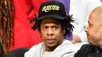 Jay-Z seated, looking contemplative, wearing a black cap with the word "RHUDE" on it.