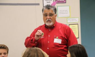 A man in a red shirt is speaking to a group of students in a classroom setting.