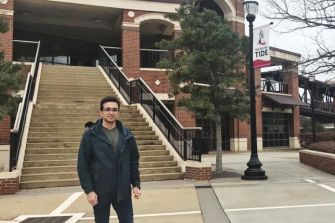 A student standing outside a university building, with steps leading up to the entrance, displaying a banner with the word "TIDE."