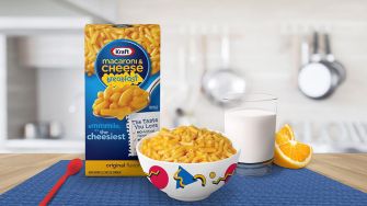 Image of a Kraft Macaroni & Cheese breakfast product, featuring a bowl of macaroni, a glass of milk, and an orange slice, against a kitchen backdrop.