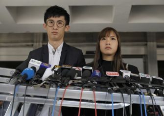 A young man and woman stand at a podium surrounded by microphones, likely preparing to address the media regarding political issues in Hong Kong.